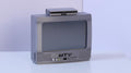 May include: A miniature silver-colored television set with a screen, a small rectangular device resting on top, and the letters MTV printed in black on the front panel. The unit features a speaker grille pattern and several small circular control buttons.