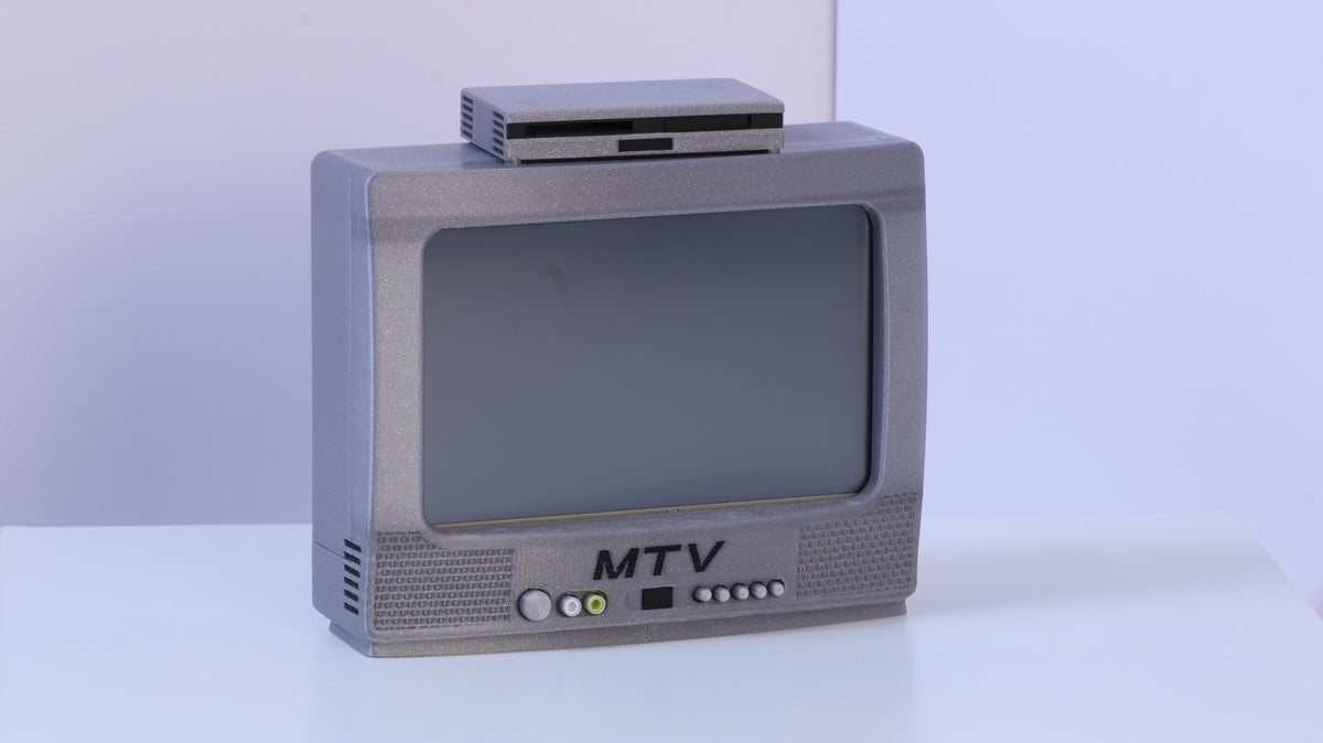 May include: A miniature silver-colored television set with a screen, a small rectangular device resting on top, and the letters MTV printed in black on the front panel. The unit features a speaker grille pattern and several small circular control buttons.