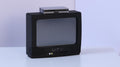 May include: A black television set with a gray screen and the letters MTV printed in white on the front panel. A small, rectangular, silver-colored electronic device is positioned on top of the television unit. The television features two circular input ports in green and white, along with a series of small control buttons.
