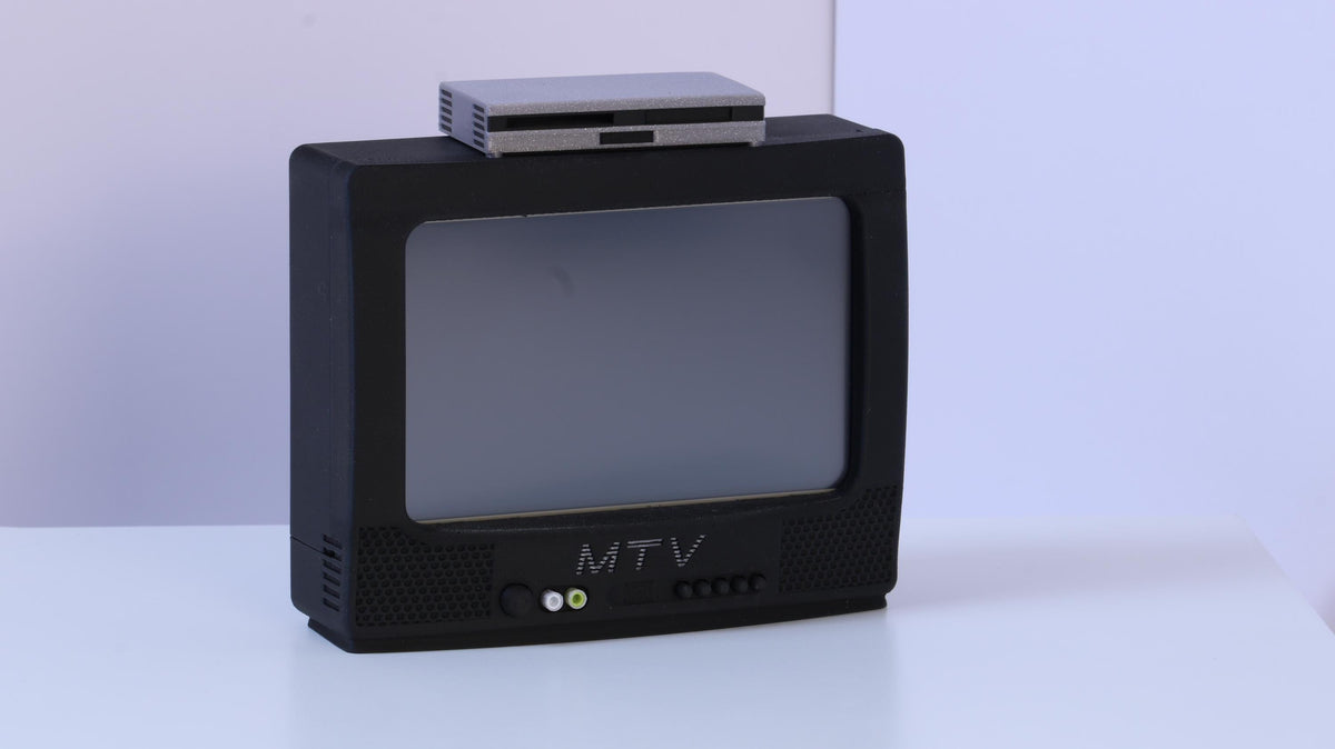 May include: A black television set with a gray screen and the letters MTV printed in white on the front panel. A small, rectangular, silver-colored electronic device is positioned on top of the television unit. The television features two circular input ports in green and white, along with a series of small control buttons.